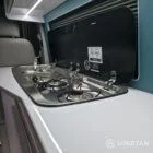 Spartan campervan galley kitchen setup with fridge, sink, and modern splashback beside passenger seats.