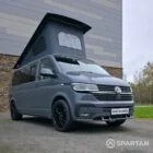 Volkswagen T6.1 long-wheelbase in Pure Grey, fitted with a Spartan camper conversion featuring the slide-and-swivel bench, clean interior lines and subtle exterior styling, parked outdoors.