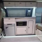 VW T6.1 interior of a Spartan campervan conversion, fitted with altro flooring, ivory and indigo blue kitchen and taupe upholstery. Featuring a microwave, fridge/ freezer, wireless fast charger, sink, double gas burner and central control panels